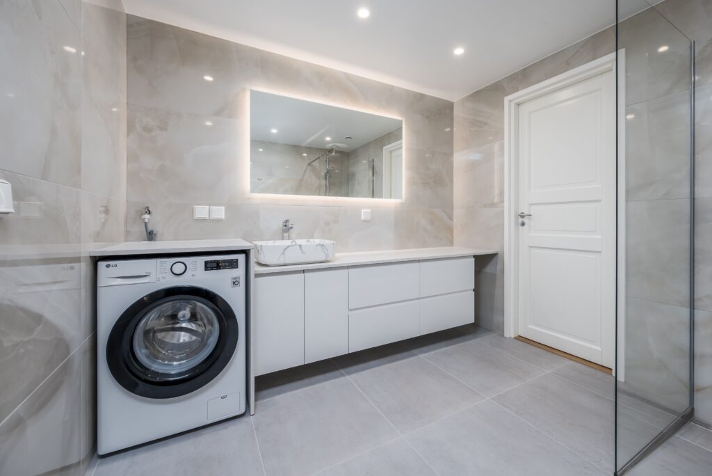 Baño moderno y luminoso con lavadora integrada, muebles blancos minimalistas y espejo retroiluminado – fotografía inmobiliaria profesional